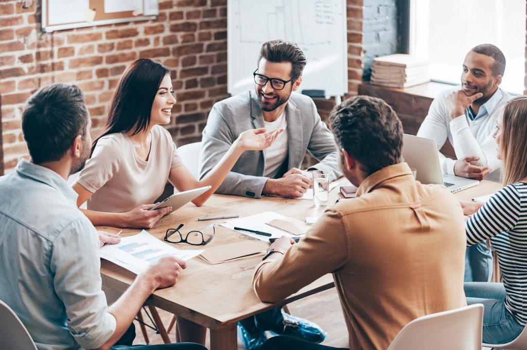 Group of smiling young professionals sitting around a wooden table at a business meeting in a modern office.